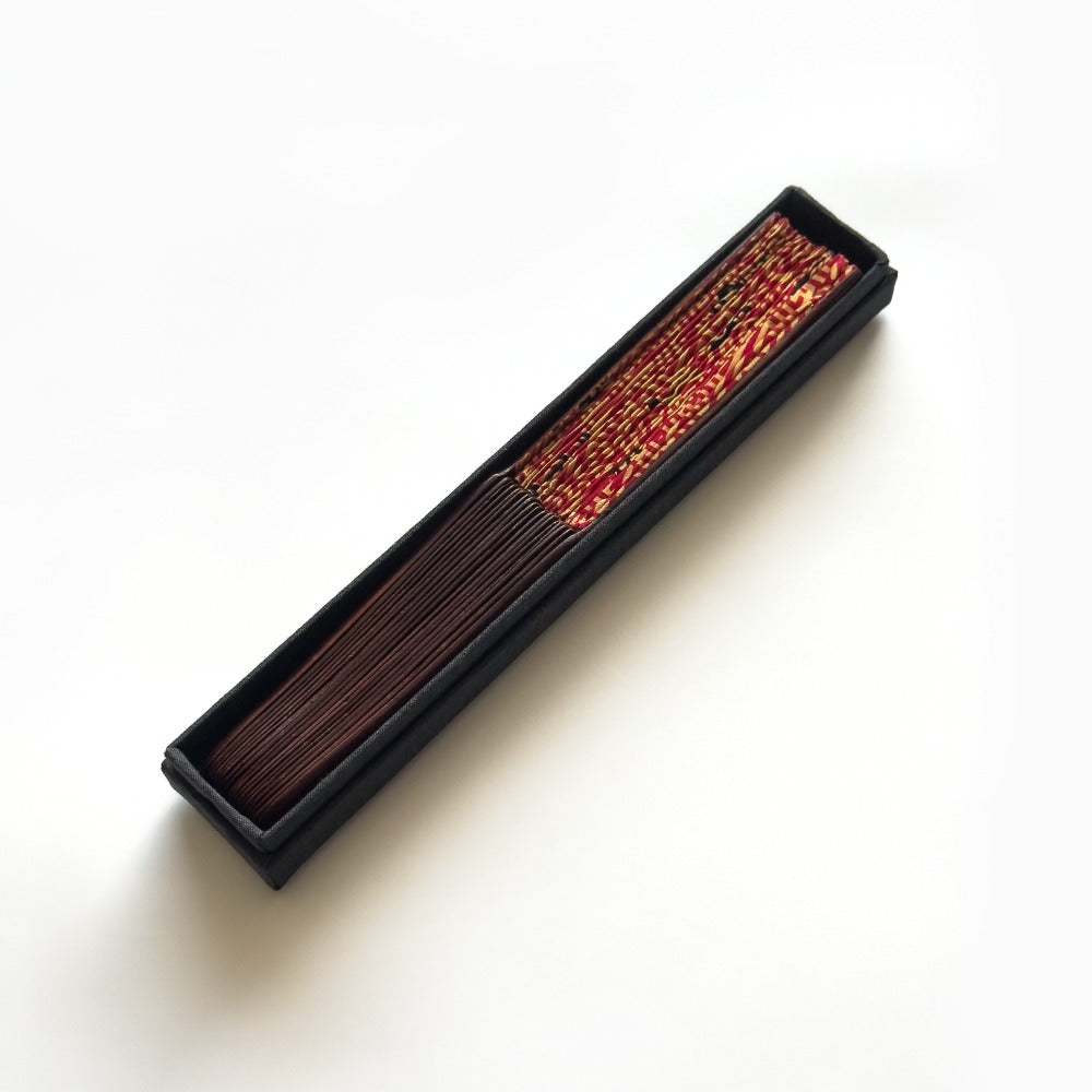 A rectangular black box containing dark brown incense sticks, with a decorative red and gold patterned cover visible at one end, photographed on a clean white background.