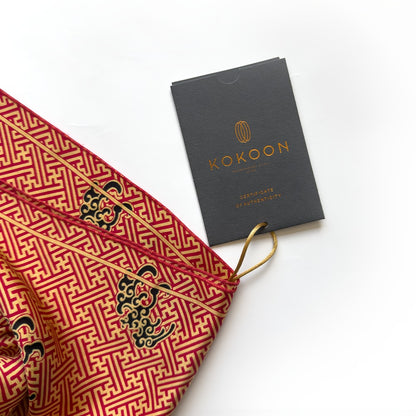 A close-up of a red and gold patterned fabric accessory with dragon motifs, shown next to a dark gray KOKOON brand tag on a white background.
