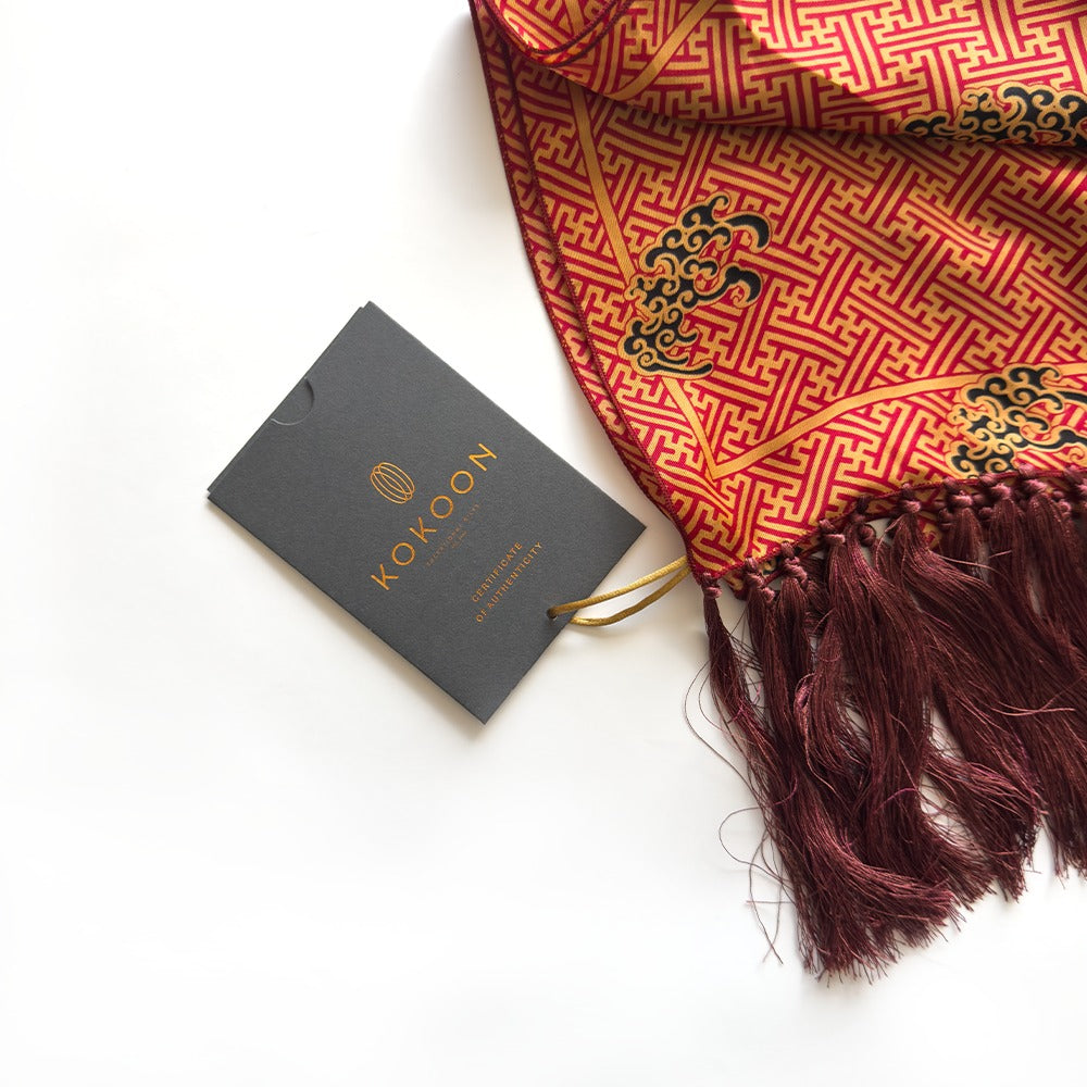 A folded red and gold patterned shawl with dark burgundy tassels, shown next to a dark gray KOKOON brand tag on a clean white background.
