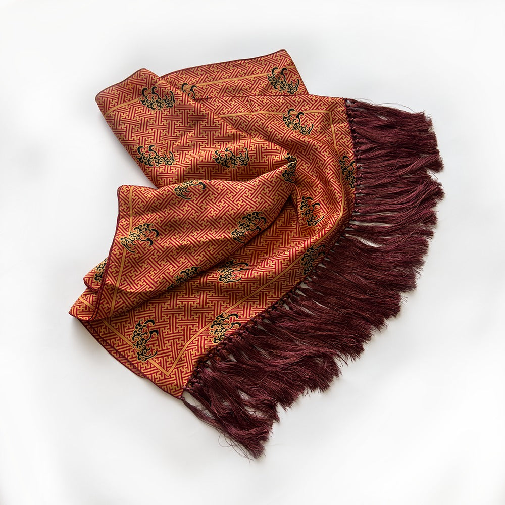 A folded patterned shawl with dark burgundy fringe, displayed on a clean white background.